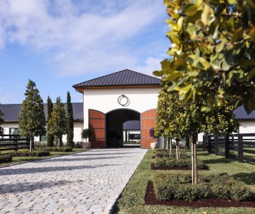 Eingang zu den 3 Graces Dressage Stallungen mit gepflastertem Weg, gepflegter Landschaftsgestaltung und symmetrisch gepflanzten Bäumen unter blauem Himmel
