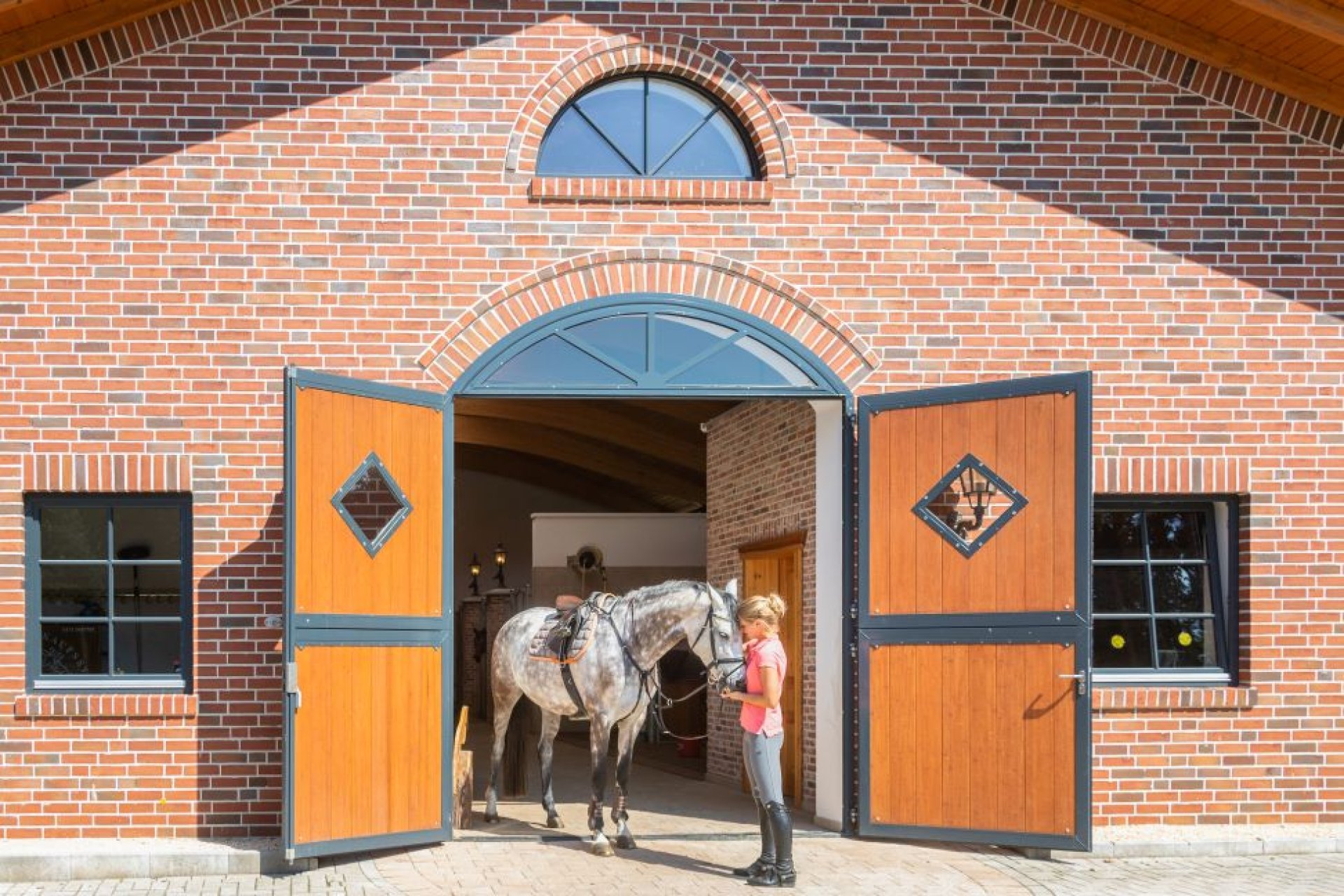 Une femme se tient avec un cheval sellé devant une porte d’écurie en bambou ouverte, avec fenêtre en losange, intégrée dans une façade en briques.