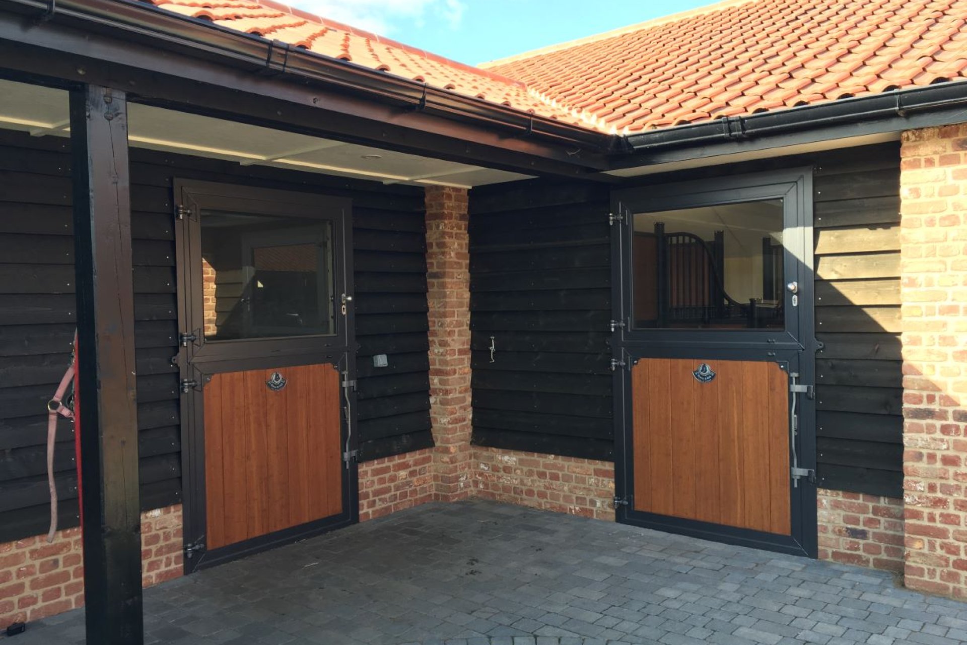 Vue extérieure d'une écurie avec des portes extérieures bicolores en anthracite et remplissage en bambou, intégrées dans une façade en bois et en brique.
