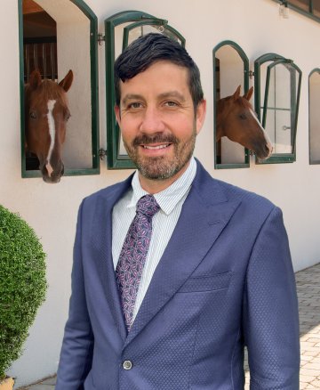 Mathew stands in front of an equestrian centre with horses looking out of the window.