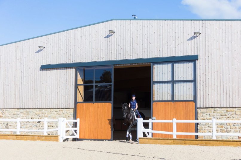 Équipement d’écurie avec grande porte coulissante en deux parties, éléments à barreaux, couvre-rail, et cavalière sortant du manège avec son cheval.
