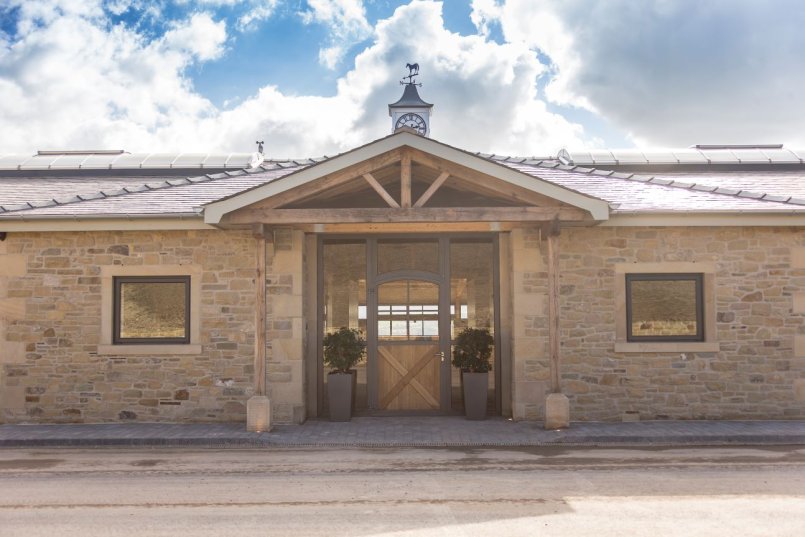 Équipement d’écurie avec entrée en pierre naturelle, grande porte d’écurie avec éléments en verre et croix en bambou, surmontée d’une horloge et d’une girouette en forme de cheval.