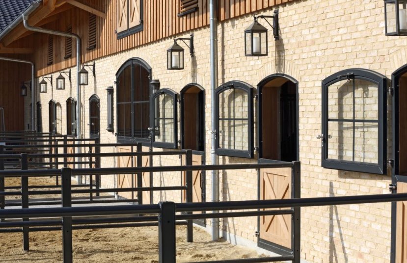 Façade d’écurie avec boxes extérieurs en briques, fenêtres cintrées anthracite et portes avec remplissage en chêne, ainsi que paddocks à l’avant.