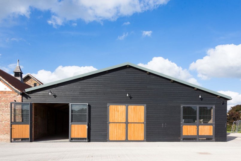 Équipement d’écurie avec une façade habillée en noir et des portes à quatre battants en verre et bois massif, alliant lumière naturelle et fonctionnalité.